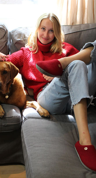 L�chelnde Frau sitzt bequem auf einer Couch, streichelt einen Hund und tr�gt rote Footprint Hausschuhe